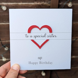 May include: A white birthday card featuring a red heart design and the text "to a special sister" and "Happy Birthday". The card is held in a hand against a wooden backdrop.