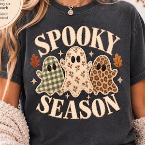 May include: A charcoal gray short-sleeve t-shirt featuring a graphic print of three ghosts with the text "SPOOKY SEASON". The ghosts have green gingham, floral, and leopard print patterns. The design includes small stars and autumn leaves.
