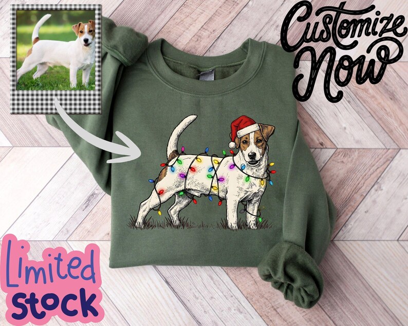 May include: Green sweatshirt featuring a Jack Russell Terrier wearing a Santa hat and Christmas lights. The sweatshirt has a photo of a Jack Russell Terrier on the upper left. Text on the image says "Customize Now" and "Limited Stock".