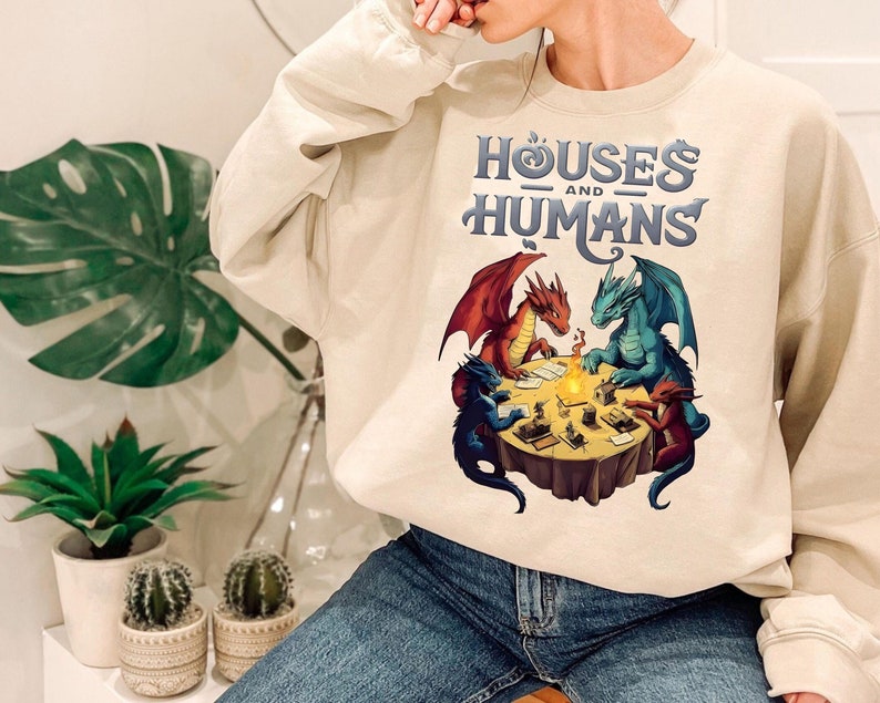 May include: A cream-colored sweatshirt with a graphic of three dragons playing a game around a table. The text "Houses and Humans" is printed on the front of the sweatshirt.