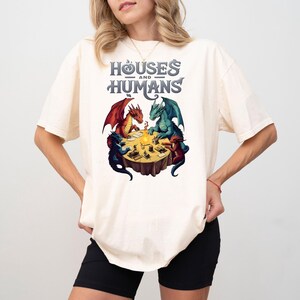May include: A white t-shirt with a graphic of three dragons playing a game around a table. The text "Houses and Humans" is above the dragons.