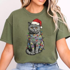 May include: Olive green t-shirt featuring a gray cat wearing a Santa hat and wrapped in colorful Christmas lights. The cat is sitting upright, with its tail curled. The design is a festive holiday theme.
