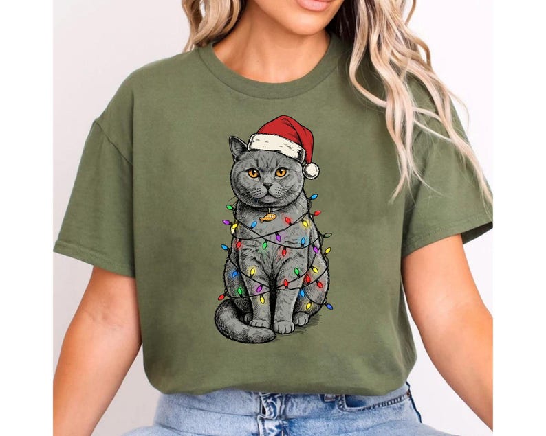 May include: Olive green t-shirt featuring a gray cat wearing a Santa hat and wrapped in colorful Christmas lights. The cat is sitting upright, with its tail curled. The design is a festive holiday theme.