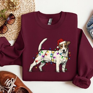May include: Burgundy sweatshirt featuring a Jack Russell Terrier wearing a Santa hat and wrapped in colorful Christmas lights. The dog is standing on a grassy area. A festive holiday-themed apparel item.