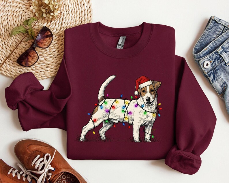 May include: Burgundy sweatshirt featuring a Jack Russell Terrier wearing a Santa hat and wrapped in colorful Christmas lights. The dog is standing on a grassy area. A festive holiday-themed apparel item.