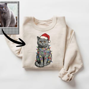 May include: A cream-colored sweatshirt featuring a graphic of a gray cat wearing a Santa hat and wrapped in colorful Christmas lights. A framed photo of a gray cat is in the upper left corner. The sweatshirt has long sleeves.