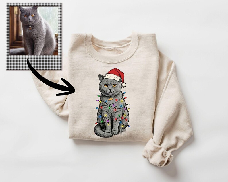 May include: A cream-colored sweatshirt featuring a graphic of a gray cat wearing a Santa hat and wrapped in colorful Christmas lights. A framed photo of a gray cat is in the upper left corner. The sweatshirt has long sleeves.