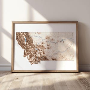Puede incluir: Un mapa en relieve enmarcado de Montana, con detalles topográficos en tonos marrones y beige. Líneas azules indican ríos y lagos. El mapa está en un marco de madera clara, contra una pared blanca.