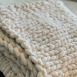 May include: A cream-colored chunky knit throw blanket with a soft, fluffy texture.