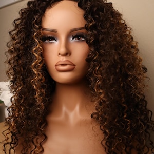 Puede incluir: Una cabeza de maniquí con una peluca rizada marrón y rubia. La peluca presenta una base marrón oscuro con reflejos rubios más claros en los rizos. El maniquí tiene una tez bronceada, labios carnosos y cejas oscuras.