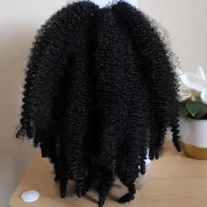 Può includere: Un'acconciatura nera con texture afro, con riccioli stretti, è esposta su una testa di manichino bianca. L'acconciatura è il punto focale, mostrando la sua consistenza e il suo volume. Lo sfondo è di un colore neutro chiaro.
