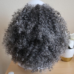 May include: A gray curly wig styled on a white wig stand. The wig has tight, voluminous curls and a natural-looking texture. The wig is displayed on a light wooden surface.