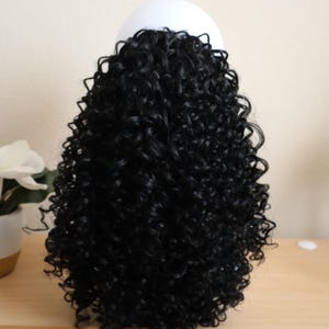 May include: A black curly wig displayed on a white mannequin head. The wig has tight, voluminous curls, giving a full and textured look. The hair is a deep, solid black, and the wig is shown on a light-coloured wooden surface.