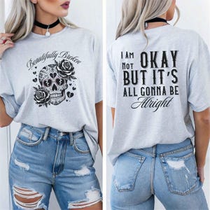 May include: Light gray t-shirt with a skull design and the words "Beautifully Broken" on the front. The skull is decorated with roses and hearts. The back of the shirt says "I am not okay but it's all gonna be alright".