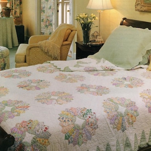 May include: A white quilt with floral wreath patterns in pastel colors, featuring a scalloped edge. The bed is in a room with a yellow armchair, a lamp, and a nightstand with a lamp and flowers.