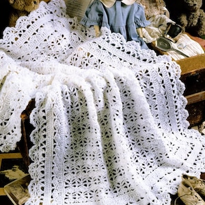 Può includere: Una coperta per neonati bianca, finemente lavorata all'uncinetto con un bordo smerlato. La coperta è drappeggiata su una cassa di legno, accanto a una bambola vintage con un vestito blu, un orsacchiotto e scarpine da neonato. Il motivo traforato della coperta è visibile.