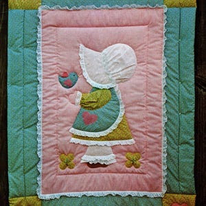 May include: A quilted applique pattern featuring a girl holding a bird. The quilt has a pink gingham center, a teal border, and yellow corner squares with heart appliques. The text "Fast, Fun Applique Pattern" is at the top.