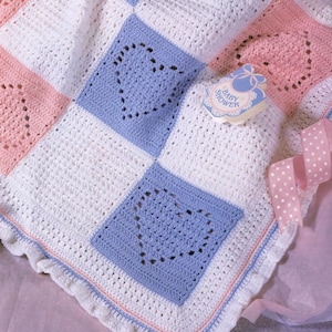 Puede incluir: Una manta de bebé de ganchillo hecha a mano en blanco, rosa y azul. La manta presenta patrones cuadrados con recortes de corazón y un borde con volantes. También se ve un recuerdo de baby shower y una cinta rosa con lunares.