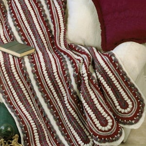 May include: A handmade crochet blanket in shades of burgundy, white, and gray, draped over a white couch. The blanket features a striped design with textured edging. A small book rests on the blanket, and a burgundy pillow is visible.