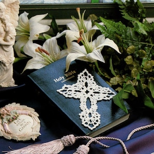May include: A still life featuring a blue Holy Bible with a white crocheted cross, white lilies, and a small angel figurine. A decorative angel-themed frame and a tassel are also present. The scene is set on a dark blue surface.