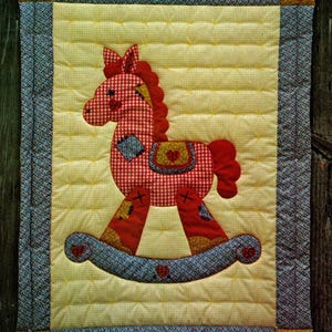 May include: A quilted wall hanging featuring a red and white checkered rocking horse applique on a yellow background. The horse has a red mane and tail. The border is blue with red hearts in the corners. The text "Fast, Fun Applique Pattern" is at the top.