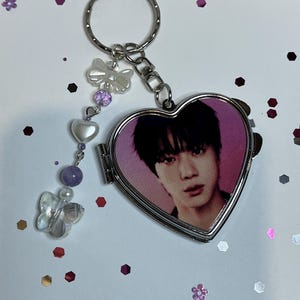 May include: A heart-shaped silver metal locket keychain featuring a portrait of a person with dark hair on the front. A dangling charm attachment includes a translucent butterfly, a white heart bead, and purple spherical beads on a silver chain.