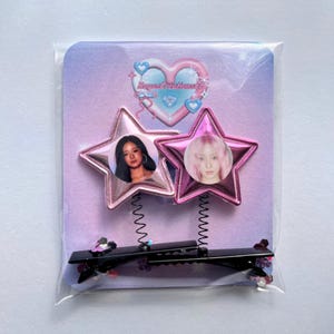 May include: Two pink star-shaped hair clips featuring images of women's faces. The clips are attached to a black barrette with a spring mechanism. The background is a light purple card with a heart and text.