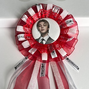 May include: A red and white rosette badge with a circular image of a person in the center. The badge features red and white ribbons, musical notes, and a decorative lace trim.