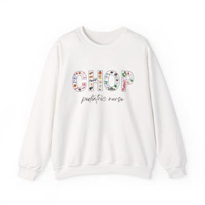 May include: White crewneck sweatshirt with the word "CHOP" in colorful letters, each filled with various small illustrations. Below the word is the text "pediatric nurse" in a handwritten style font. A comfortable, casual top for healthcare professionals.