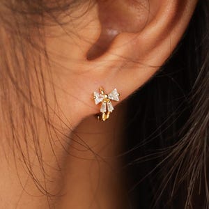 May include: A gold-coloured stud earring featuring a bow design, embellished with small, clear gemstones. The earring has a delicate and elegant appearance. The bow is the focal point, with the gemstones adding sparkle.