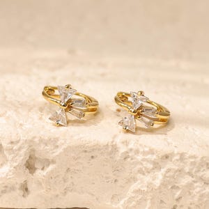 May include: Two gold-tone hoop earrings, each adorned with a cluster of clear, geometric crystals. The earrings are presented on a textured, off-white surface, highlighting their design and sparkle.