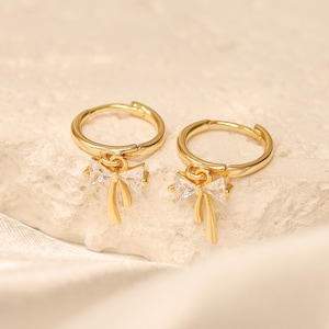 Può includere: Un paio di orecchini a cerchio color oro. Ogni orecchino presenta una piccola pietra trasparente a taglio rettangolare e un accento pendente color oro. Gli orecchini sono esposti su una superficie chiara e strutturata.