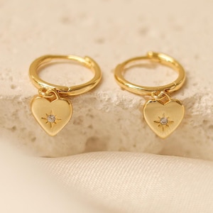 May include: Gold hoop earrings with a small heart charm featuring a sparkling rhinestone in the center.