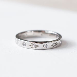 Puede incluir: Un anillo de banda de plata con una fila de pequeñas piedras transparentes y grabados en forma de estrella.