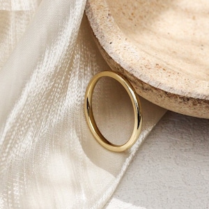 Può includere: Un semplice anello a fascia d'oro su uno sfondo bianco con un tessuto beige drappeggiato dietro.