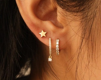 May include: A close-up of a person's ear with three gold earrings. The top earring is a small star, the middle earring is a small hoop with clear stones, and the bottom earring is a dangling earring with a small clear stone.