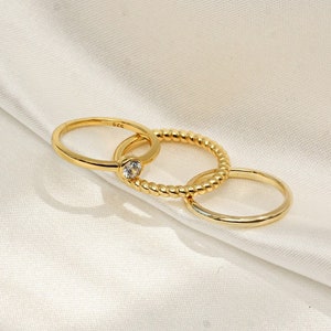 May include: Three gold rings, one with a small clear stone, stacked on a white fabric background.