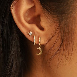 May include: A person's ear with three gold earrings. The top earring is a small star with a single clear stone in the center. The middle earring is a small hoop with clear stones set in the metal. The bottom earring is a hoop with a crescent moon charm hanging from it.