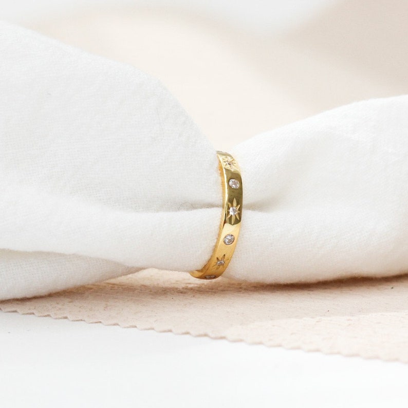 K&ouml;nnte beinhalten: Ein goldener Bandring mit einer Reihe kleiner, klarer Edelsteine und sternf&ouml;rmigen Akzenten.