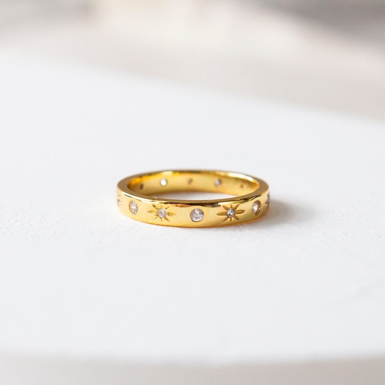 K&ouml;nnte beinhalten: Ein goldener Bandring mit einem zarten Design aus kleinen Sternen und klaren Edelsteinen.
