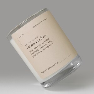 May include: A clear glass candle jar with a beige label. The label reads "impossible" with the text "your forever is valid, (darling, remember), you are unstoppable." The candle is white, and the scent is "stonefruit + amber."