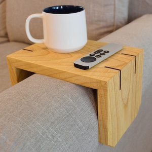 Puede incluir: Una bandeja de brazo de sofá de madera clara con una taza y un mando a distancia. La bandeja es rectangular con detalles en marrón oscuro en las esquinas. Una taza blanca con interior azul oscuro está sobre la bandeja, junto a un mando a distancia plateado.