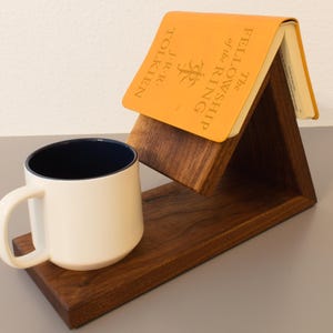 May include: A dark brown wooden book stand with a triangular shape holds an orange book titled The Fellowship of the Ring by J.R.R. Tolkien. A white ceramic mug with a dark blue interior sits on the flat base of the wooden stand.