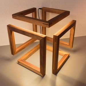 Puede incluir: Una lámpara de madera moderna con un diseño geométrico. La lámpara presenta una estructura en forma de cubo compuesta por marcos triangulares interconectados. La madera tiene un tono cálido y natural, y se ve un cable de alimentación negro. La lámpara está encendida, proyectando sombras suaves.