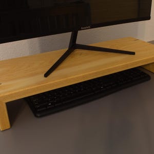 May include: A light-colored wooden monitor stand with a black keyboard underneath. The stand is rectangular with a raised platform and a black monitor on top. The stand is made of light-colored wood and has a simple, modern design.