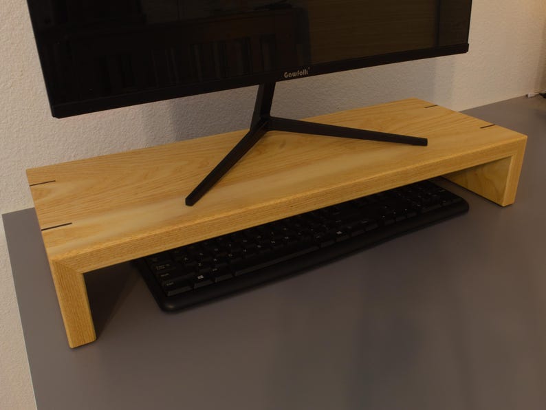 May include: A light-colored wooden monitor stand with a black keyboard underneath. The stand is rectangular with a raised platform and a black monitor on top. The stand is made of light-colored wood and has a simple, modern design.