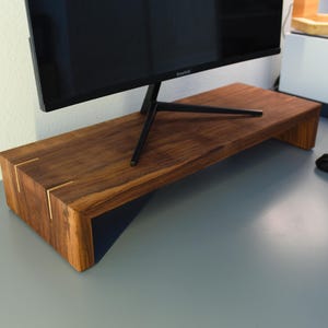 May include: A wooden monitor stand with a dark brown finish. The stand has a rectangular shape with a black support underneath. The corners are accented with gold-colored inlays. A computer monitor sits on top of the stand.