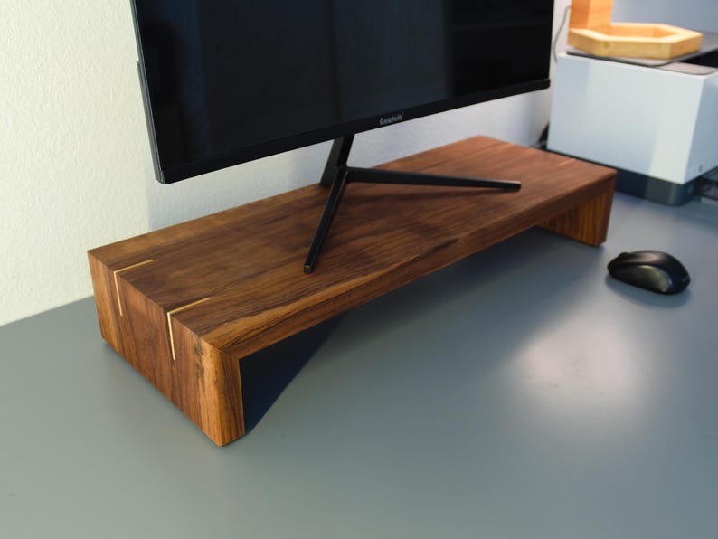 May include: A wooden monitor stand with a dark brown finish. The stand has a rectangular shape with a black support underneath. The corners are accented with gold-colored inlays. A computer monitor sits on top of the stand.