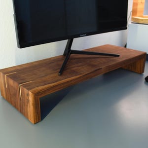 May include: A dark brown wooden monitor stand with a rectangular shape and a black monitor on top. The stand has gold accents on the corners and is designed to elevate a computer monitor. A black computer mouse is on the desk.
