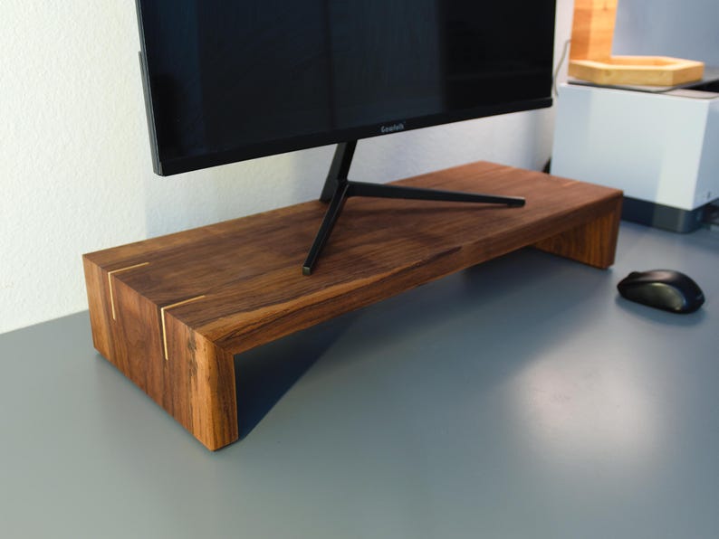 May include: A dark brown wooden monitor stand with a rectangular shape and a black monitor on top. The stand has gold accents on the corners and is designed to elevate a computer monitor. A black computer mouse is on the desk.